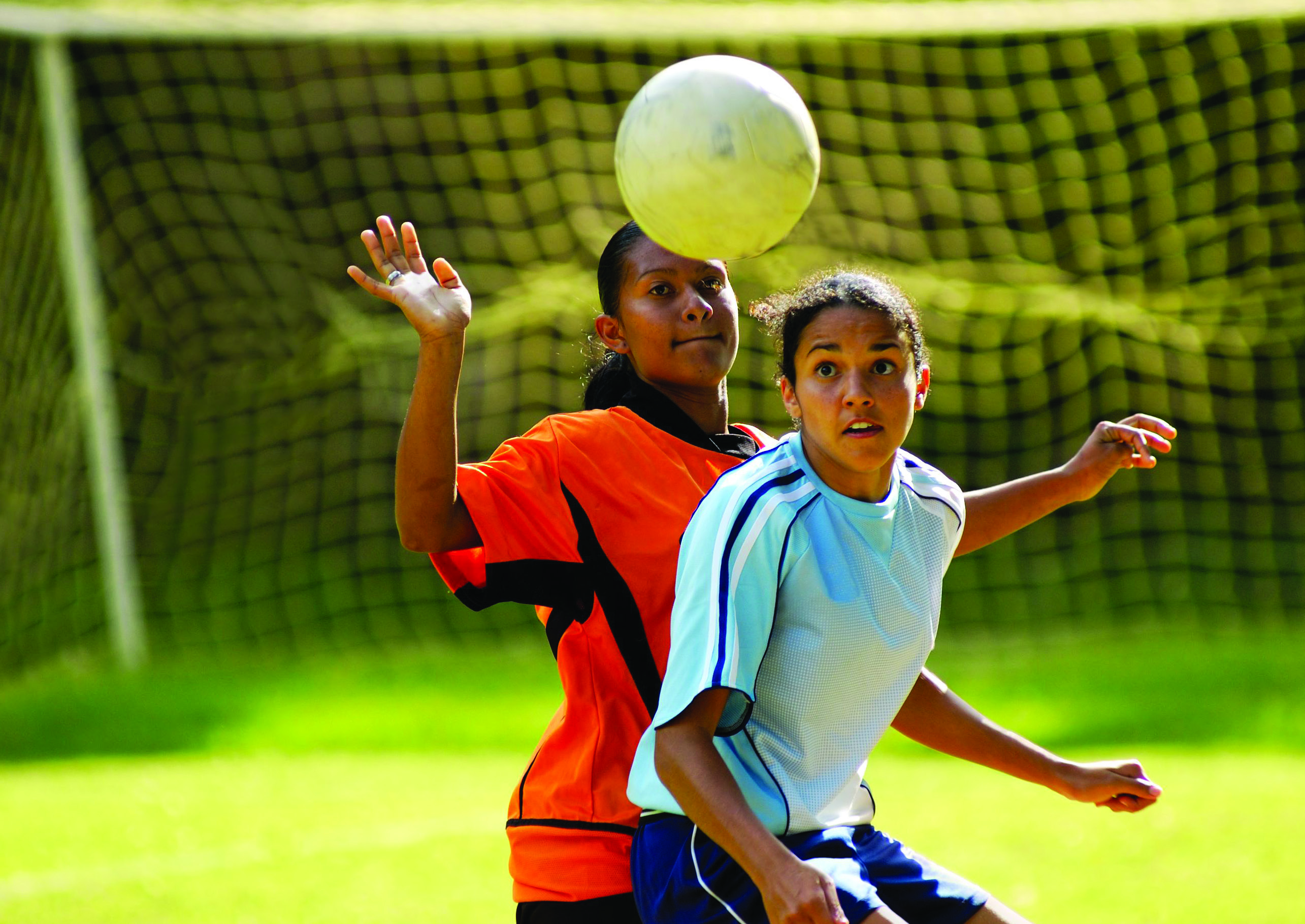 Two teenage girls are playing soccer on opposing teams as a ball flies toward the goal net.