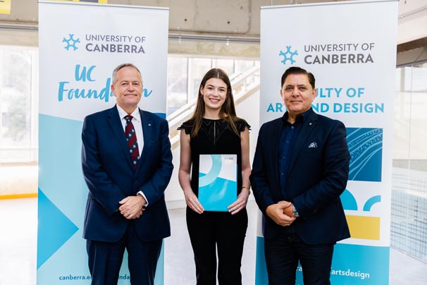 Grace Careless (middle) holding her scholarship certificate, with Vice-Chancellor Bill Shorten on the left and UC Council Member Medy Hassan on the right standing in front of University of Canberra banners