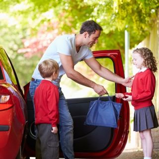 A father dropping two children off at school, handing his daughter a school backpack.