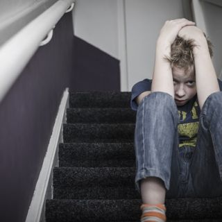 A scared looking young boy with his head in his hands, sits on a flight of stairs