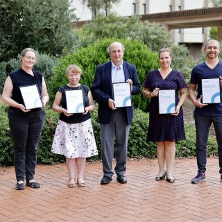 Five of the award winners and representatives standing, holding their awards against a background of green plants