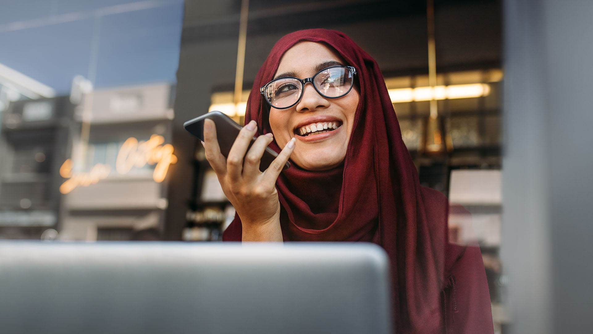 A UC student wearing glasses and smiling in a dark red hijab holding a smartphone